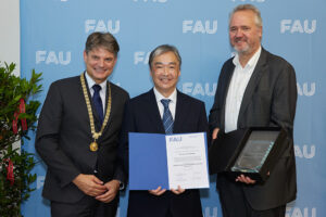 3 people in dark suits stand in front of a light blue wall on which FAU is written in white bush letters. The three people look smilingly into the camera, the person in the middle holds a blue folder with a white paper inside in front of his body.