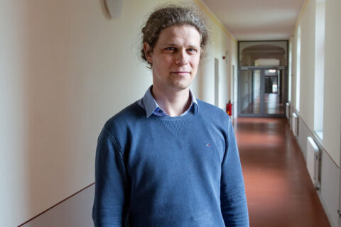 A person with a blue sweater and gray-blond hair looks into the camera. The person is standing in a hallway of a building. The floor is red and a glass door can be seen in the background.