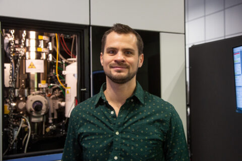 Portrait photo of Prof. Pelz. Prof. Pelz has short brown hair and a beard; he is standing in front of a window through which a machine can be seen.