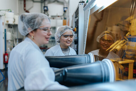 Two people in white coats, hairnets and safety goggles stand in front of a machine behind a yellowish pane of glass. Two black hoses lead through the glass pane.