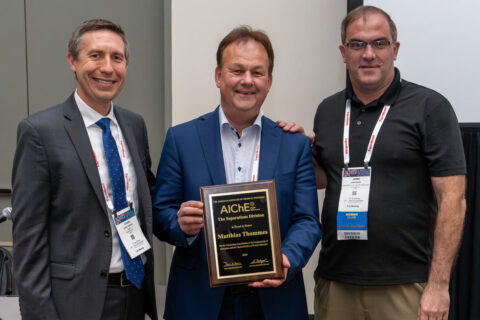 3 people stand next to each other and smile friendly into the camera. The person in the middle is holding a certificate in front of their body.